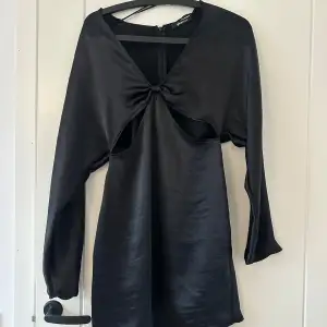 Snygg svart klänning från Gina Tricot i storlek XS. Den har en unik design med en knutdetalj framtill och långa ärmar. Perfekt för en utekväll eller fest! Materialet är mjukt och glansigt, vilket ger en elegant look. Den är oanvänd men har tvättats en gång.