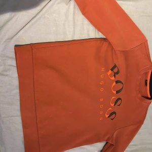 Orange tröja från Hugo Boss - Säljer en snygg orange röd tröja från Hugo Boss. Den har en stor logga på framsidan och är perfekt för en avslappnad stil. Tröjan är i bra skick och passar perfekt för höst och vår. Den har långa ärmar och en bekväm passform.