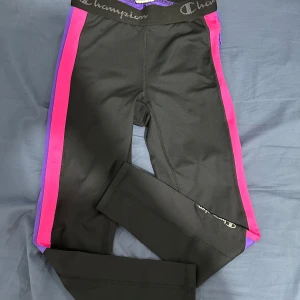Svarta leggings från Champion - Säljer ett par svarta leggings från Champion i storlek S. De har snygga rosa och lila detaljer längs sidorna och äett elastiskt midjeband med Champion-loggan. Perfekta för träning eller en avslappnad dag. Materialet är mjukt och stretchigt för bästa komfort.