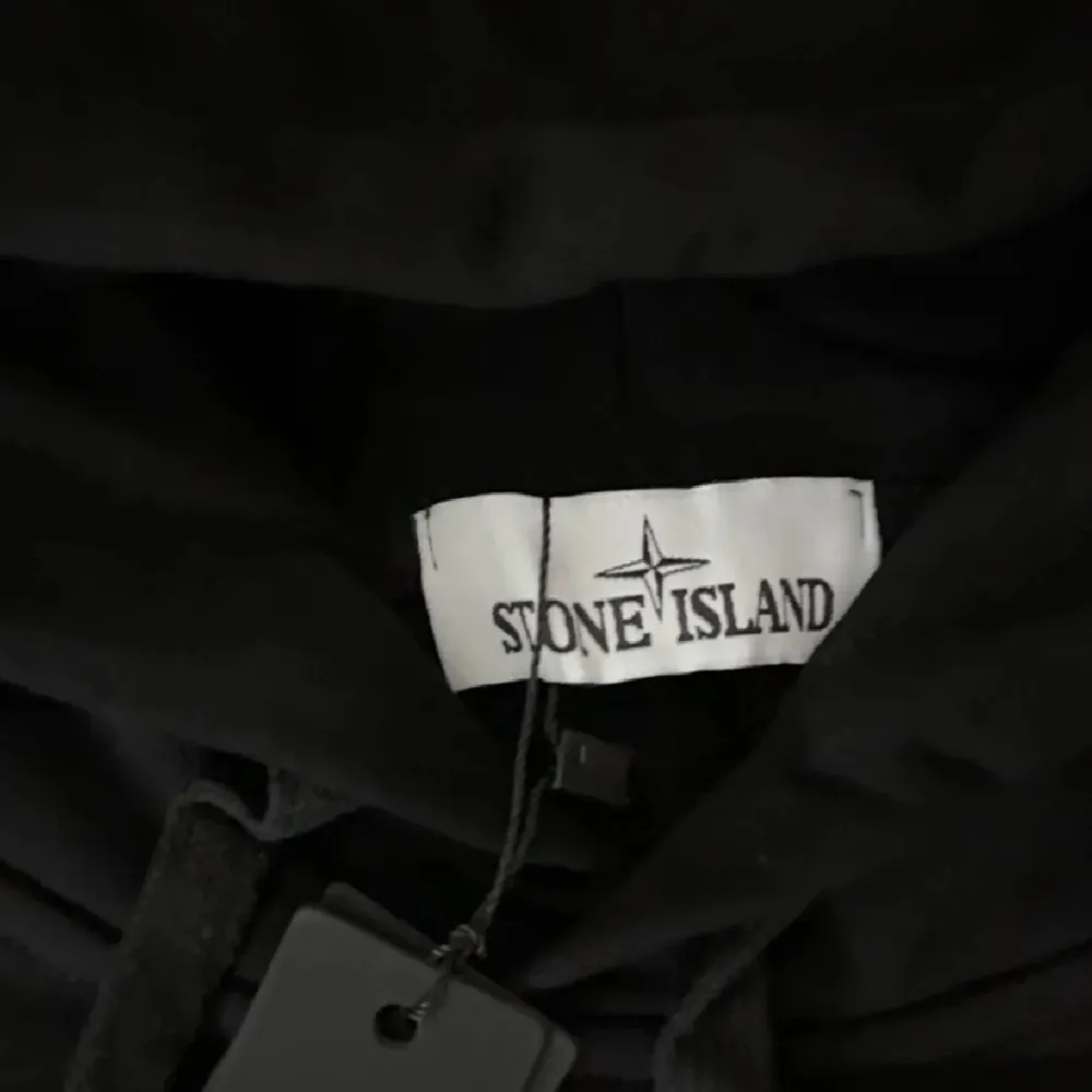 Säljer en snygg svart hoodie från Stone Island. Den har en klassisk design med huva och en ikonisk logga på ärmen. Perfekt för en casual look eller att bara mysa i. Helt ny med taggar kvar!. Tröjor & Koftor.