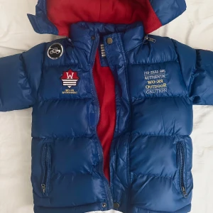 Blå pufferjacka från Knot so Bad - Säljer en blå pufferjacka från Knot so Bad i storlek 110 (5 år). Jackan har en mysig röd fleecefoder och är dekorerad med broderade märken på framsidan. Den är helt ny och knappt använd. Perfekt för kalla dagar med sin varma och vadderade design. Den har en dragkedja framtill och en praktisk huva. Passar perfekt för vinter och höst! 