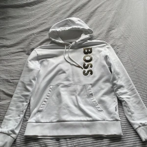 Vit hoodie från Boss - Säljer en snygg vit hoodie från Boss i ny skick. Den har en stor logga på bröstet och en praktisk magficka. Perfekt för en avslappnad stil och passar till alla säsonger. Kvitto finns självklart med! Butik pris 2000kr