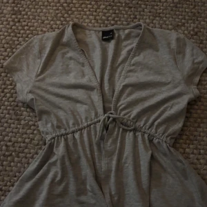 Grå topp från Gina Tricot - Säljer en enkel och stilren grå topp från Gina Tricot i storlek XS. Toppen har korta ärmar och en knytning i midjan för en smickrande passform. Perfekt för vardagsbruk eller en avslappnad dag ute. Materialet är mjukt och bekvämt.