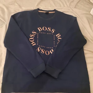  Hugo Boss tröja  - Säljer en snygg mörkblå sweatshirt från Hugo Boss med ett coolt tryck i guld rosa på framsidan. Tröjan har en normal passform och är perfekt för både höst och vår. Den är i mycket bra skick och superbekväm att ha på sig. Perfekt för en casual look!
