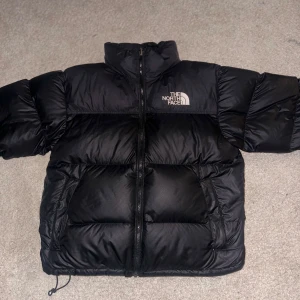 Svart dunjacka från The North Face - Säljer en svart dunjacka från The North Face i mycket bra skick. Jackan har en pufferstil med dragkedja och är perfekt för kalla vinterdagar. Den är långärmad och har justerbar dragsko i nederkant för extra värme.   