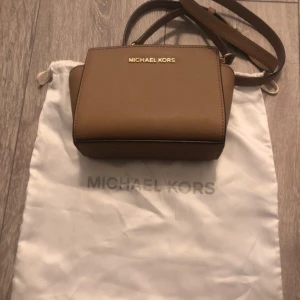 Brun axelremsväska  - Säljer en snygg brun axelremsväska med gulddetaljer från Michael Kors, som är i väldigt bra skick. Dock finns en liten fläck på undersidan av väskan men inget som syns när man väl använder (bild 4). Perfekt för både vardag och fest. Kommer med dustbag!