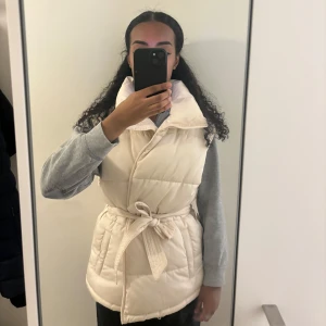 Beige dunväst med bälte - Säljer en snygg beige dunväst i mycket bra skick. Västen har en praktisk dragkedja och ett bälte i midjan för en figursydd look. 
