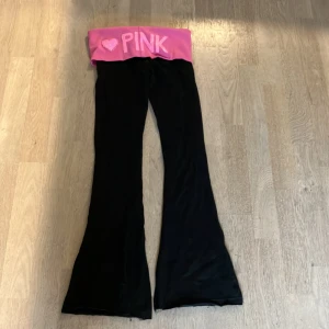 Svarta byxor med rosa midja  - Säljer ett par svarta byxor med en bred rosa midja . De har en hjärtdetalj och texten 'PINK' i glittriga bokstäver. Byxorna är i bootcut-stil och ser superbekväma ut för en avslappnad dag hemma eller på stan.