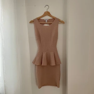 Beige klänning från NLY One - Snygg beige klänning från NLY One i storlek XS. Den har en peplumdetalj i midjan och en öppen rygg med korsade band. Perfekt för festliga tillfällen! Materialet är stretchigt och bekvämt.