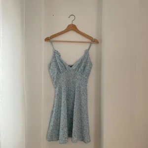 Blommig sommarklänning - Söt ljusblå klänning med blommigt mönster, perfekt för sommardagar. Den har smala axelband och en v-ringad design som ger en feminin touch. Klänningen är i ett lätt och luftigt material, vilket gör den idealisk för varma dagar. Passar bra till både vardag och festliga tillfällen.