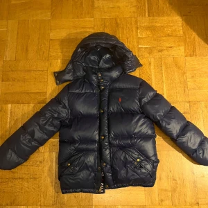 164/ XL i barnstorlek. Mörkblå dunjacka från Polo Ralph Lauren - Säljer en snygg mörkblå dunjacka från Polo Ralph Lauren. Den har en röd insida och en praktisk huva. Jackan är perfekt för kalla dagar och har både dragkedja och knappar framtill. Den är i mycket bra skick och redo för vintern.