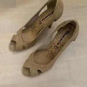 Beige pumps från Tamaris - Snygga beige pumps från Tamaris med öppen tå och hög klack. Tillverkade i läder och ett textilliknande material!