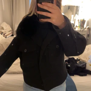 Svart kappa med pälsdetalj - Snygg svart kappa med pälsdetalj på kragen. Jackan har knappar framtill och två fickor. Är i äkta päls. Pris går att diskutera, inga skambud. 