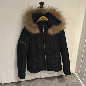 Svart jacka med pälsdetalj från Attentif  - Snygg svart jacka från Attentif med quiltade detaljer och en mysig pälsfodrad huva (avtagbar). Jackan har en dragkedja framtill och praktiska fickor med dragkedjor. Perfekt för kyliga dagar! Uppskattad storlek 38. 
