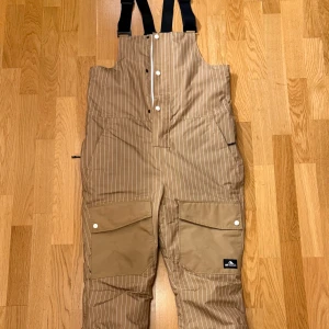 Snowboardbyxor från Warp - Snygga täckbyxor från Warp med vita ränder och justerbara hängslen. Byxorna har praktiska fickor med knappar framtill och en dragkedja baktill. Perfekta för goa åk i backen👌