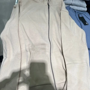 Beige  hoodie från - Säljer en stilren beige hoodie från . Den har en dragkedja framtill och en praktisk ficka på magen. Perfekt för lager-på-lager eller som ett lättare plagg under varmare dagar.