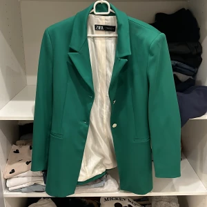 Grön kavaj från Zara - Snygg grön kavaj från Zara med klassisk design och vita knappar. Perfekt för att ge en färgklick till din outfit. Kavajen har en stilren passform och är tillverkad i ett mjukt material som ger en bekväm känsla.