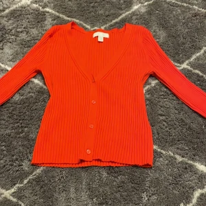 Röd ribbad kofta från H&M - Snygg röd kofta från H&M med ribbad struktur och v-ringning. Den har långa ärmar och knappar framtill, perfekt för att lägga till en färgklick i garderoben. Passar bra till både jeans och kjol.