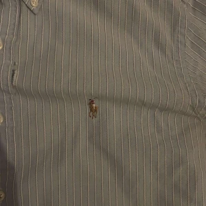 Randig skjorta från Ralph Lauren - Säljer en klassisk randig skjorta från Ralph Lauren i storlek L. Skjortan har en broderad logotyp på bröstet och knappar framtill. Perfekt för en stilren look. Har en defekt på framsidan!