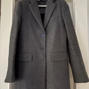 Klassisk grå kappa från Zara - Snygg grå kappa i ull från Zara med klassisk design och tre knappar framtill. Den har långa ärmar och två fickor med lock. Fodrad med ett elegant svart tyg för extra komfort.