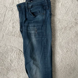Blå jeans med slitningar - Snygga blå jeans med en cool slitning på framsidan. De har en klassisk femficksdesign och en knappgylf. Perfekta för en avslappnad stil.