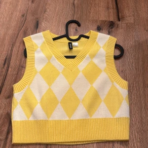 Gul och vit stickad väst - Snygg gul och vit stickad väst från H&M med ett klassiskt harlekinmönster. Västen har en v-ringad design och ribbade kanter som ger en stilren look. Perfekt för att lägga till lite färg i din garderob!