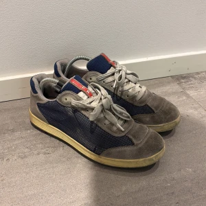 Grå och blå sneakers Prada  - Snygga sneakers i grått och blått med snörning och en röd detalj på plösen. Skorna har en robust sula och är tillverkade i mocka och syntetmaterial. Perfekta för en avslappnad stil. Märket är Prada. Skicket 9/10 ser bättre ut i värklighet en på bild. Storlek 38/halv passar 39 stora i storlek.