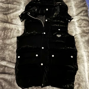 Svart dunväst från Philipp Plein - Snygg svart dunväst från Philipp Plein med blanka detaljer och dragkedja. Västen har en cool design med knappar och en logotyp på bröstet. Perfekt för att hålla stilen när det är kyligt ute. Skick 10/10 inte använd storlek S 