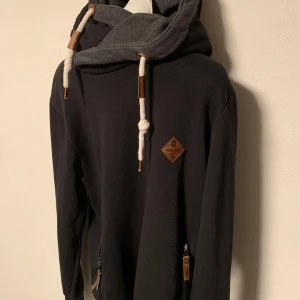 Svart hoodie med dragkedja och snörning från Indicode  - Snygg svart hoodie med grå insida i huvan och vita snören. Den har dragkedjor på fickorna och en läderdetalj på bröstet. Perfekt för en avslappnad stil. Storlek S.