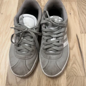 Grå Adidas sneakers - Säljer ett par grå Adidas sneakers med vita detaljer och snörning. tillverkade i mocka. Ganska smutsiga så pris går att diskutera. 