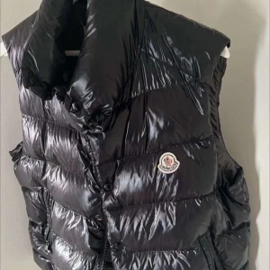 Svart dunväst från Moncler - Snygg och glansig svart dunväst från Moncler med hög krage och knäppning framtill. Perfekt för kyligare dagar när du vill hålla stilen. Monclers logga syns tydligt på bröstet.