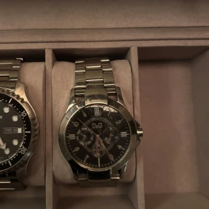 Silverfärgad klocka från D&G - Snygg silverfärgad klocka från D&G med svart urtavla och flera små urtavlor för extra funktioner. Armbandet är i metall och har en klassisk design. Perfekt för den som vill ha en stilren accessoar. Klockan är gammal men går fortfarande och använda, finns defekter (smått vattenskadad därav priset) (repor) 