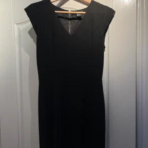 Svart klänning från H&M - Elegant svart klänning från H&M med en stilren v-ringning och korta ärmar. Klänningen har en dragkedja i ryggen för enkel på- och avtagning. 