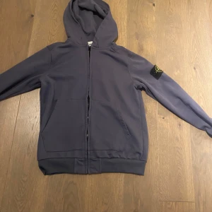 Blå huvtröja från Stone Island - Säljer en snygg blå huvtröja från Stone Island med dragkedja och huva. Tröjan har en ikonisk logga på ärmen och är perfekt för en casual look. Den har långa ärmar och ribbade muddar för extra komfort. Knappt använd 