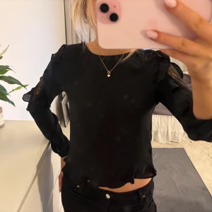Svart blus med spetsdetaljer - Snygg svart blus med spetsdetaljer på ärmarna och volanger. Blusen har långa ärmar och knappar i ryggen. Perfekt för en stilren look.