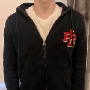 Svart stickad hoodie med dragkedja - Säljer en svart stickad hoodie från Ralph lauren med dragkedja och röd broderad Ralph lauren  logga på bröstet. Passar dig som är 170-180 ☺️☺️