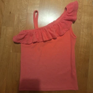 Rosafärgad oneshoulder topp - Snygg rosafärgad oneshoulder topp med volangdetalj. Perfekt för sommarens varma dagar. Toppen har en smal axelrem och är ribbad för en bekväm passform. Färgen på första bilden är ungefär hur den ser ur I verkligheten