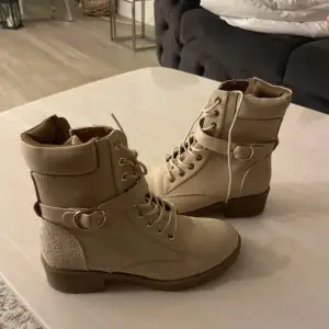 Snygga beige boots med snörning och dragkedja. De har en dekorativ rem med spänne och en robust sula. Perfekta för en stilren look.
