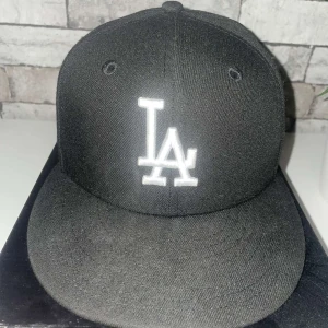 Svart keps från New Era - Snygg svart keps från New Era med vit broderad LA-logga på framsidan. Kepsen har en klassisk design med platt skärm och är perfekt för en stilren look. Märkeslogga på sidan ger en extra touch.