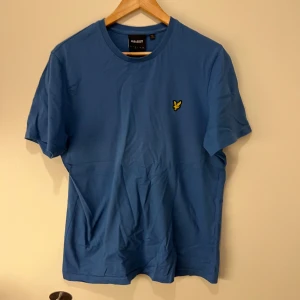 Blå t-shirt från Lyle & Scott - Säljer en snygg blå t-shirt från Lyle & Scott med korta ärmar och ett broderat gult emblem på bröstet. Den är i ett bra skick samt inget tecken på slitage! Skriv i dm om du vill diskutera!