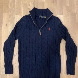 Mörkblå stickad tröja från Polo Ralph Lauren - Säljer en snygg mörkblå stickad tröja från Polo Ralph Lauren med dragkedja och röd broderad logga på bröstet. Tröjan har en klassisk kabelstickad design och långa ärmar. Perfekt för kyligare dagar! Kan diskutera om priset. Passar dig för som har storlek S-M