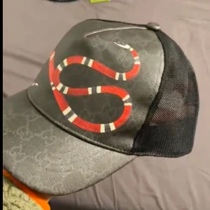 Snake keps Gucci  - Snygg svart keps med ett iögonfallande ormmönster i rött och vitt på framsidan. Kepsen har en nätpanel på baksidan för bättre ventilation och en justerbar rem för perfekt passform.