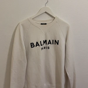 Balmain Paris - Säljer en stilren vit sweatshirt från Balmain med svart text på bröstet. Tröjan har långa ärmar och en klassisk rund halsringning. Perfekt för en avslappnad och trendig look. 