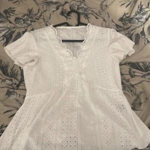 Vit blus med spetsdetaljer - Charmig vit blus med korta ärmar och v-ringning. Blusen har vackra spetsdetaljer och ett hålmönster som ger en romantisk känsla. Perfekt för varma dagar eller sommarens festligheter. Använd 1 gång 