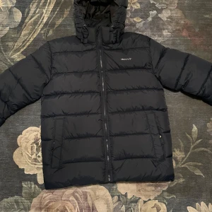 Svart pufferjacka från GANT - Säljer en svart pufferjacka från GANT med dragkedja och huva. Jackan är quiltad och har en stilren design med GANT-logga på bröstet. Perfekt för kyligare dagar. Jackan är helt oanvänd.