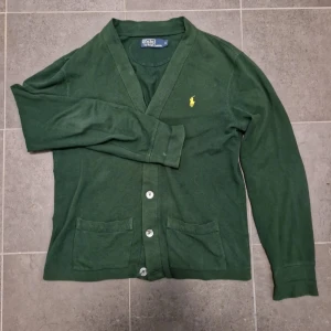 Polo Ralph Lauren kofta/cardigan - Snygg grön kofta från Polo Ralph Lauren med knappar framtill och två praktiska fickor. Den har en klassisk design med långärmade ärmar och en liten gul logotyp på bröstet. Perfekt för en stilren look. SKRIV VID FUNDERINGAR OCH🔵INNAN DU KÖPER🔵