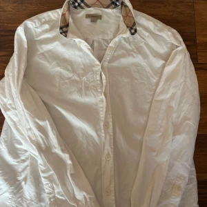 Vit skjorta från Burberry - Snygg vit skjorta från Burberry med klassisk rutig krage i beige, röd och svart. Skjortan har långa ärmar och knappar framtill. Perfekt för en stilren look.