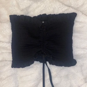 Svart smockad tubtopp från bikbok  - Snygg svart tubtopp med smockad design och volangdetaljer. Toppen har en knytning framtill som ger en justerbar passform.