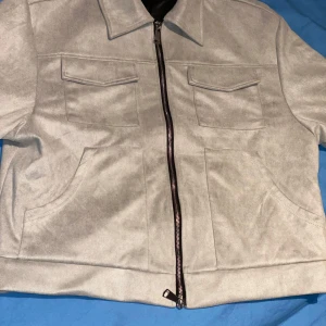 Grå mockajacka med dragkedja - Snygg grå mockajacka med dragkedja framtill och två bröstfickor med lock. Jackan har en klassisk krage och är perfekt för en stilren look. Passar bra till både vardag och mer uppklädda tillfällen.