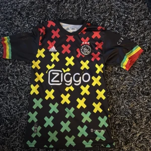 Svart fotbollströja från Adidas med färgglada kryss och Bobby på baksidan - Säljer en unik svart fotbollströja från Adidas med färgglada kryss i rött, gult och grönt. Tröjan har korta ärmar med matchande färgdetaljer och ett tryck på baksidan. Perfekt för fotbollsfans som vill sticka ut!